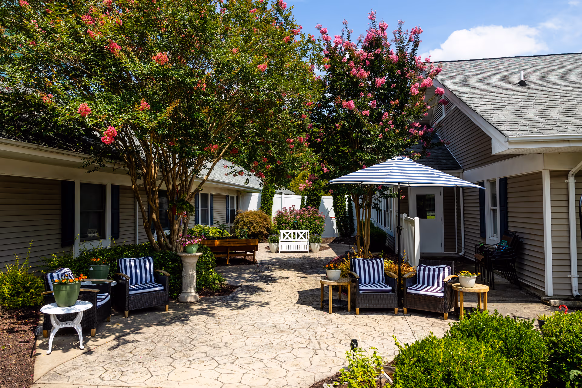 Outdoor courtyard area at Churchland Nursing Home with paved stone flooring, several seating arrangements including chairs with blue and white striped cushions, a white umbrella, potted plants, blooming pink flowering trees, and beige buildings with white trim surrounding the space.