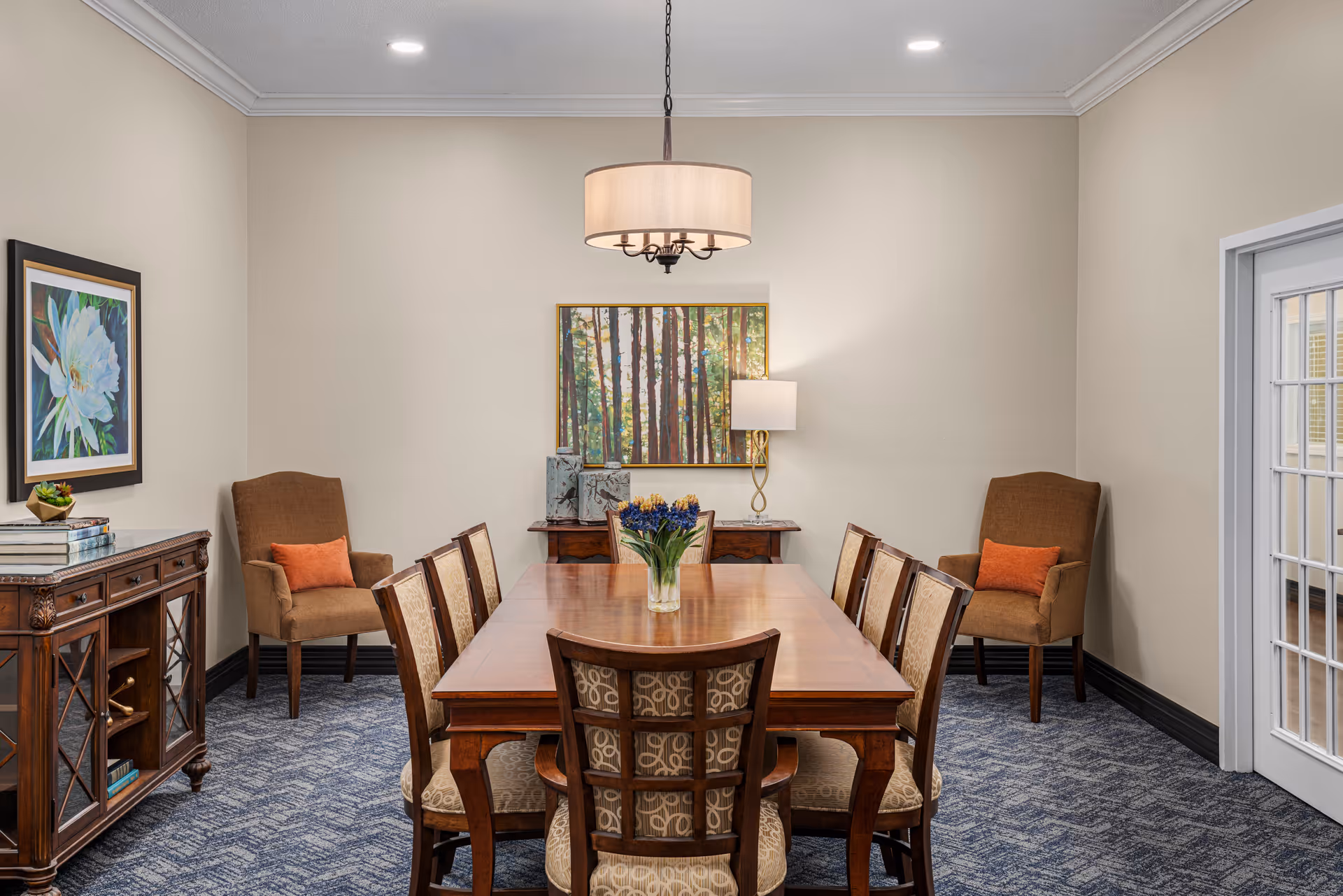 A dining room with a wooden rectangular table surrounded by eight upholstered chairs. Two brown armchairs with orange cushions are placed against the walls on either side of the room. The room has beige walls, a blue patterned carpet, and is decorated with framed artwork and a table lamp on a sideboard. A modern chandelier hangs above the table, and there is a glass-paneled door on the right side.