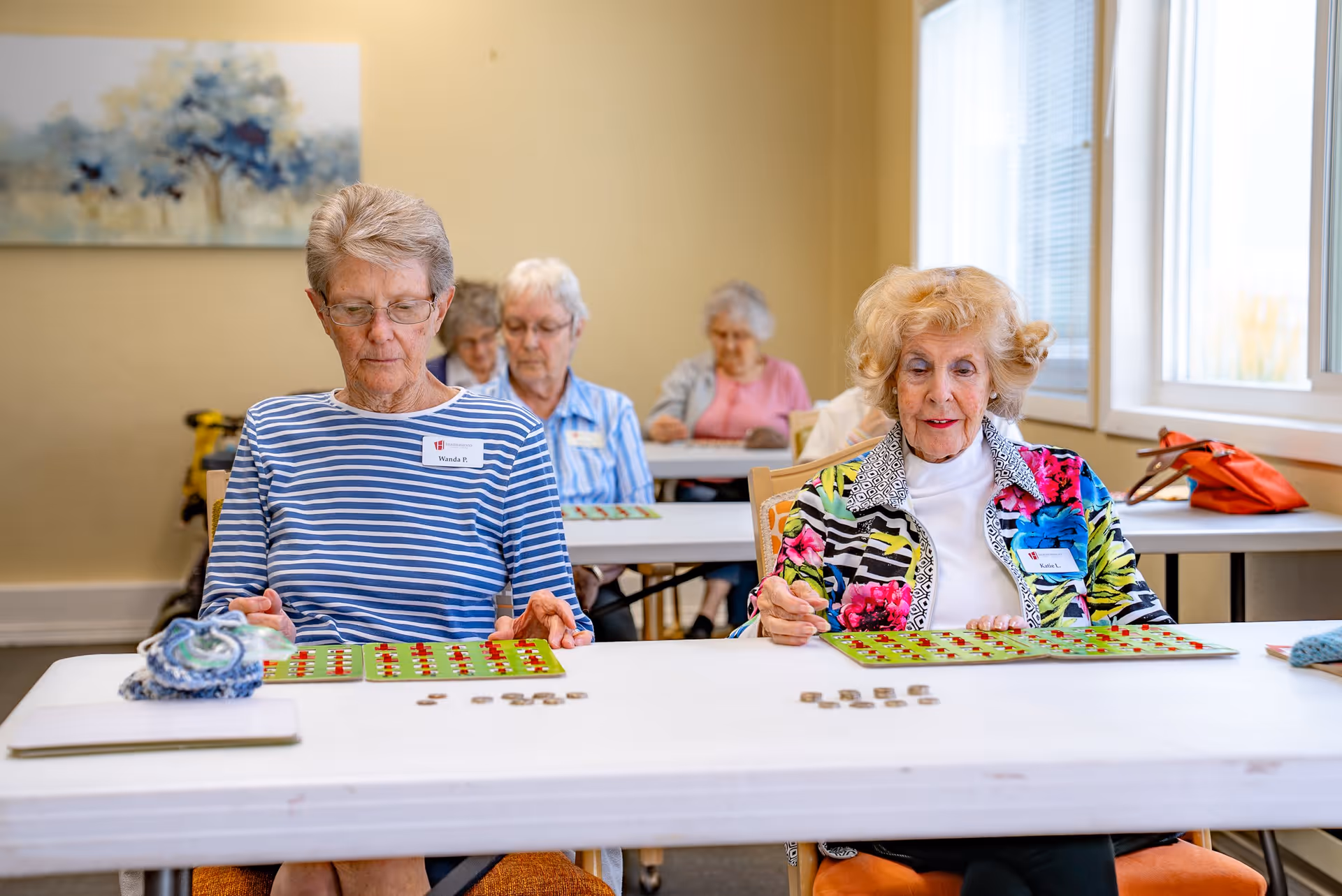 Several elderly women sitting at tables in a well-lit room playing bingo, with bingo cards and chips in front of them. The room has light-colored walls and large windows letting in natural light.