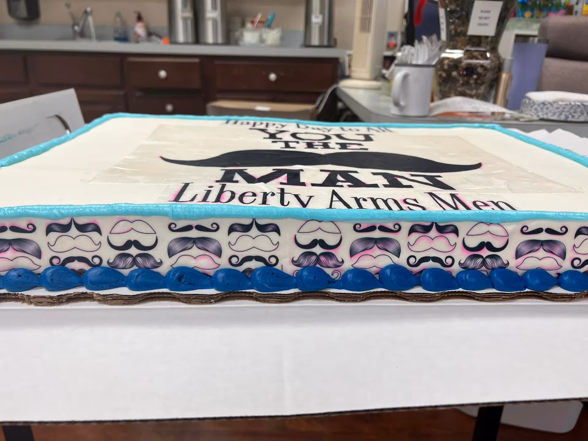 A decorated rectangular cake with a mustache design and the text 'Happy Father's Day to the MATT Liberty Arms Men' on top. The cake has a blue and white border with various mustache patterns around the sides. The background shows a kitchen or dining area with cabinets, a sink, and some utensils.