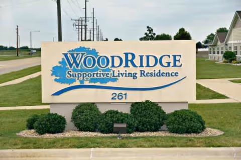Exterior sign reading WoodRidge Supportive Living Residence with greenery and a sidewalk.