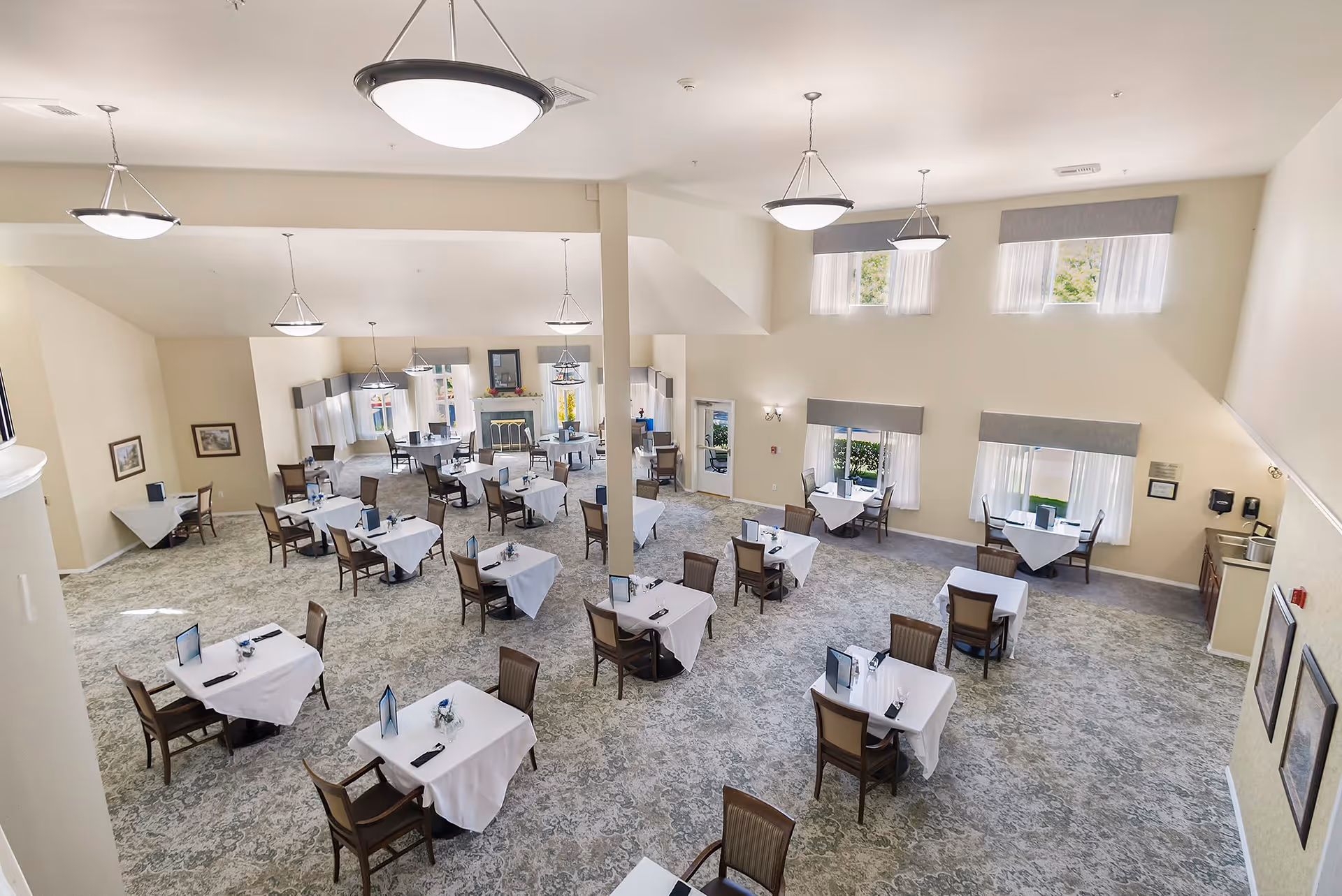 A spacious dining room with multiple tables covered in white tablecloths, each set with chairs and small centerpieces. The room has large windows with sheer curtains allowing natural light, beige walls, and carpeted floors. Several ceiling lights hang from the high ceiling, and framed pictures decorate the walls.