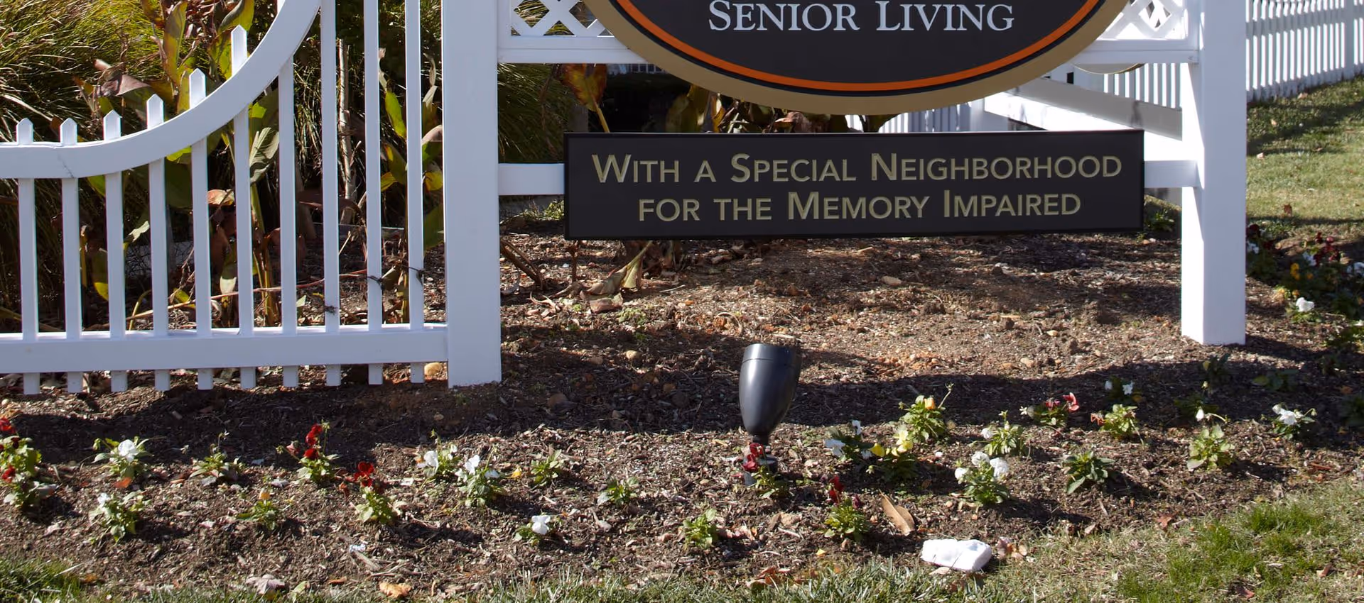 Entrance landscaping with a white picket fence and a senior living sign reading "With a Special Neighborhood for the Memory Impaired."