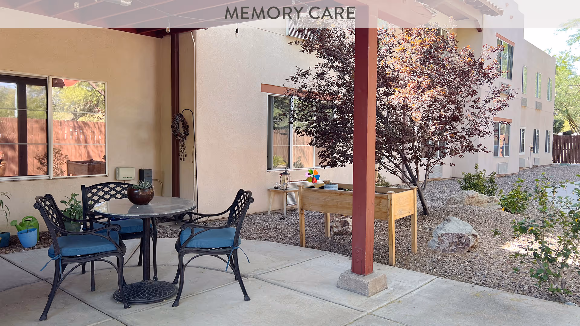 Outdoor covered patio area with a round table and four metal chairs with blue cushions. There are potted plants along the wall, a raised garden bed, and a tree with red leaves. The building exterior is visible with windows and a gravel landscape.