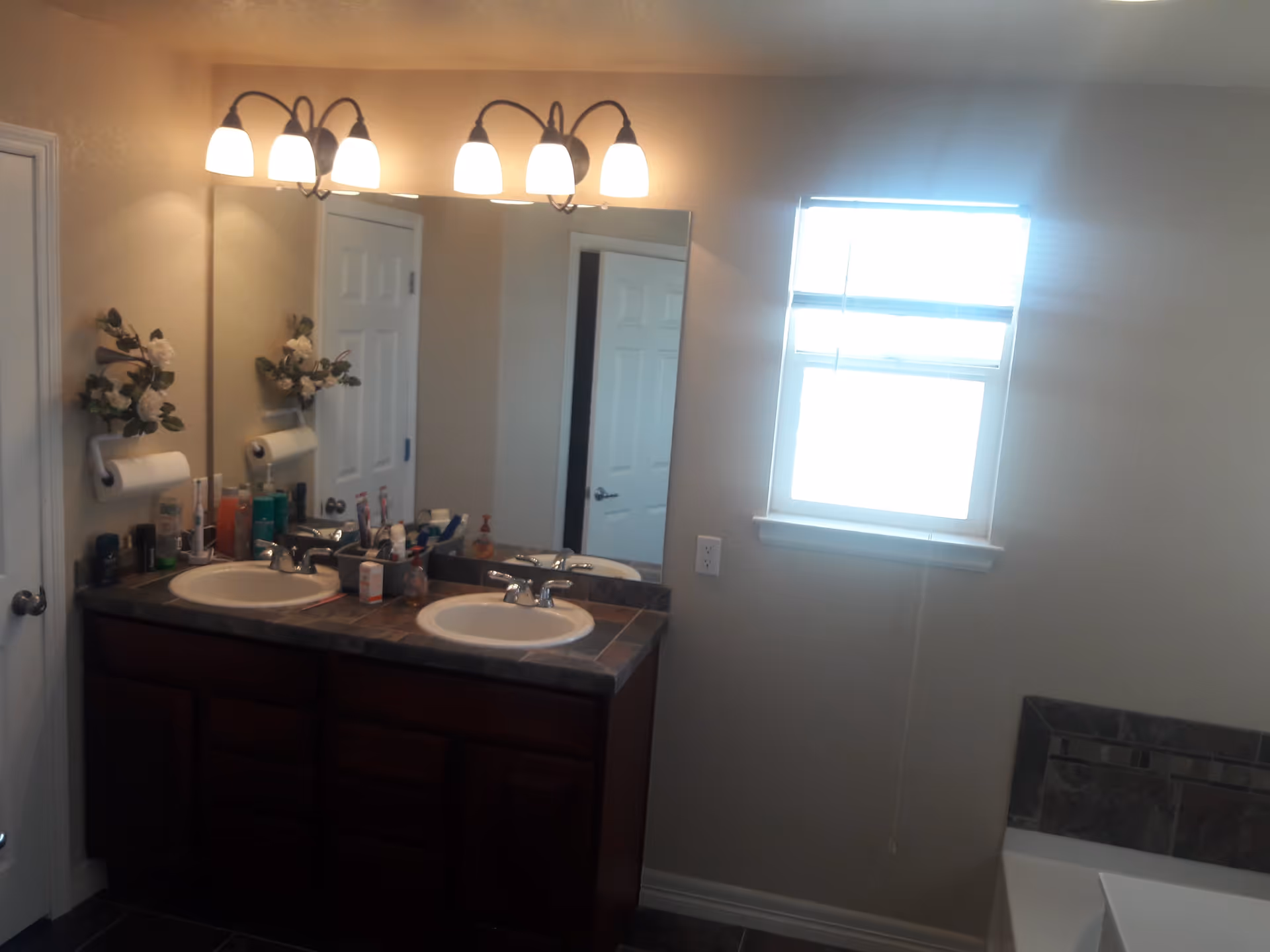 Bathroom interior with a double sink vanity featuring dark wood cabinets and a tiled countertop. Above the sinks is a large mirror with two sets of three-light fixtures. A window with blinds is on the right wall, and a bathtub with tiled surround is partially visible in the lower right corner. Various toiletries are arranged on the countertop.
