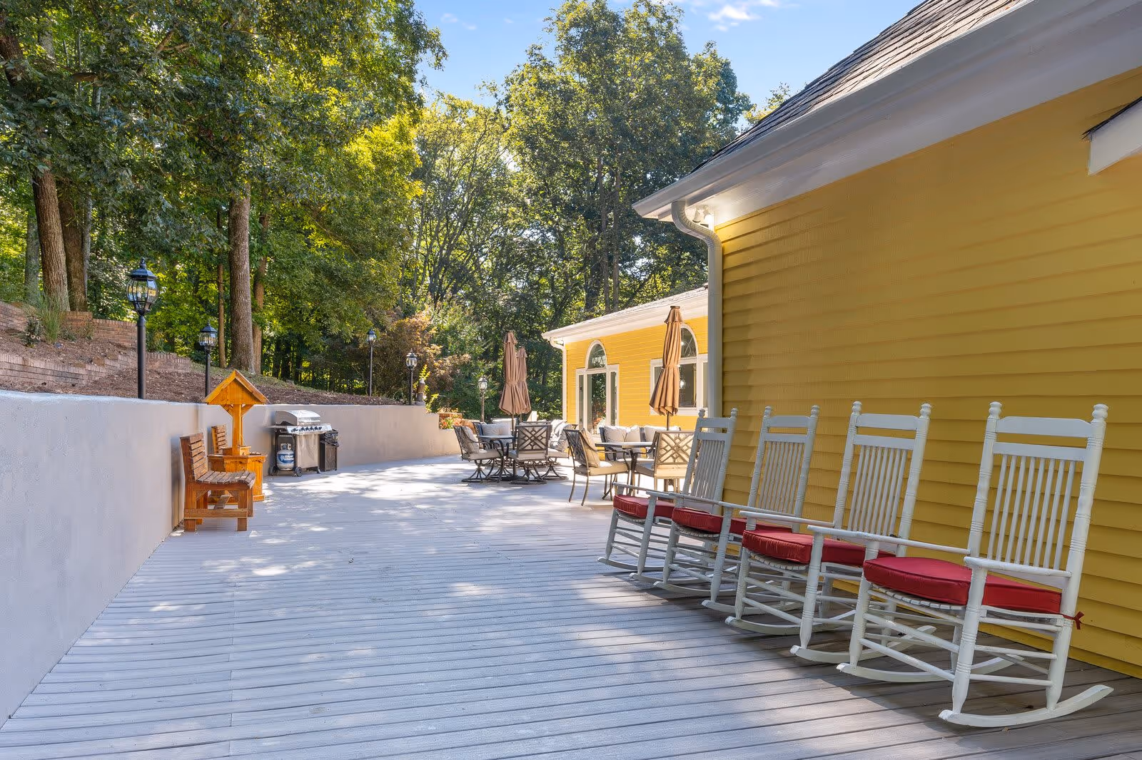 Outdoor patio area at New Hope Senior Living of Hendersonville featuring a row of white rocking chairs with red cushions along a yellow building. There are tables with umbrellas and chairs further down the patio, surrounded by trees and greenery under a clear blue sky.