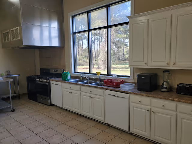 A kitchen with white cabinets and drawers, a large window overlooking trees, a double sink with dish soap and a red dish rack, a black stove with a stainless steel hood, a dishwasher, a blender, a toaster, and a small black appliance on the countertop.