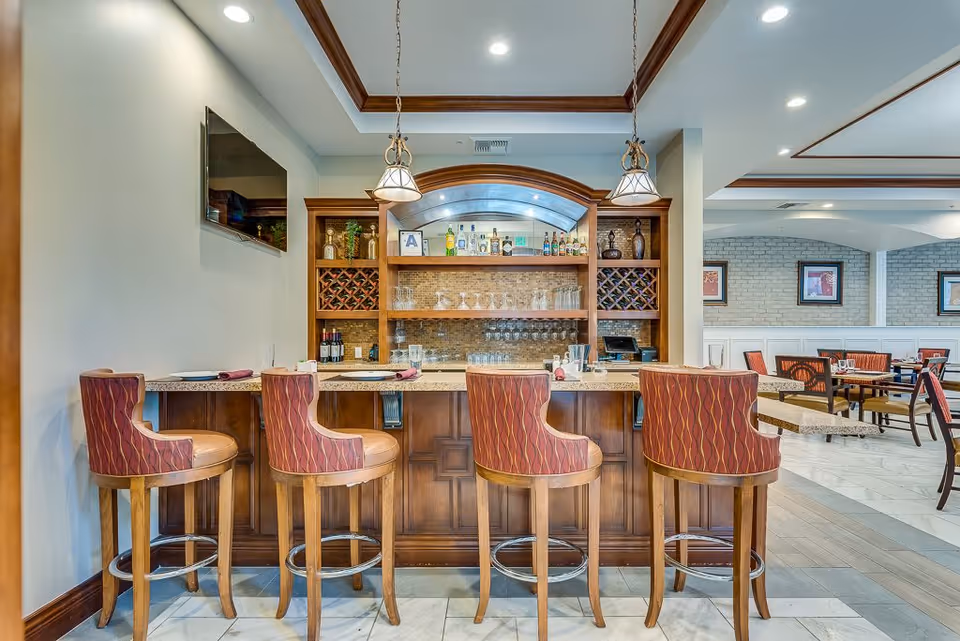 Interior view of a bar area in a senior living facility with four high wooden chairs with red patterned upholstery lined up at a granite countertop. Behind the bar are shelves stocked with various bottles of liquor, wine racks, and glassware. The room has warm lighting with two hanging pendant lights above the bar and a flat-screen TV mounted on the wall to the left. In the background, there is a dining area with tables and chairs, framed artwork on the walls, and a light-colored brick accent wall.