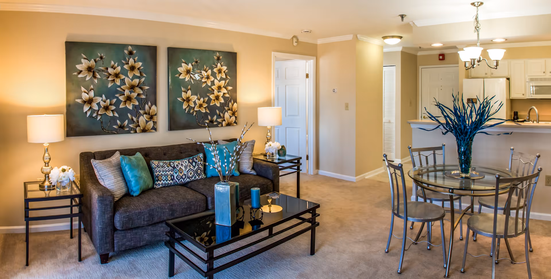 A cozy living and dining area in a senior living facility featuring a dark gray sofa with decorative pillows, two floral wall art pieces above the sofa, two side tables with lamps and flowers, a glass coffee table with blue decorative items, and a round glass dining table with four metal chairs and a blue plant centerpiece. The background shows a kitchen area with white cabinets and appliances.