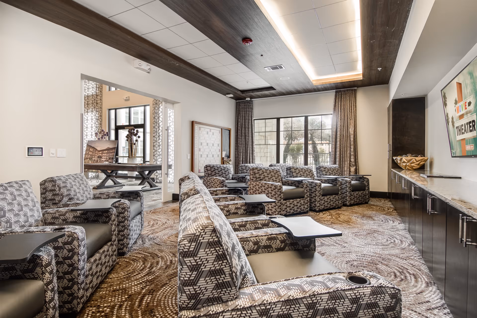 A modern theater room with multiple patterned armchairs arranged in rows, each equipped with a small attached table. The room has a large window with curtains, a patterned carpet, and a flat-screen TV mounted on the wall displaying a 'Movie Theater' sign. There is a long cabinet beneath the TV and a decorative bowl on top. The ceiling has recessed lighting with wooden accents.