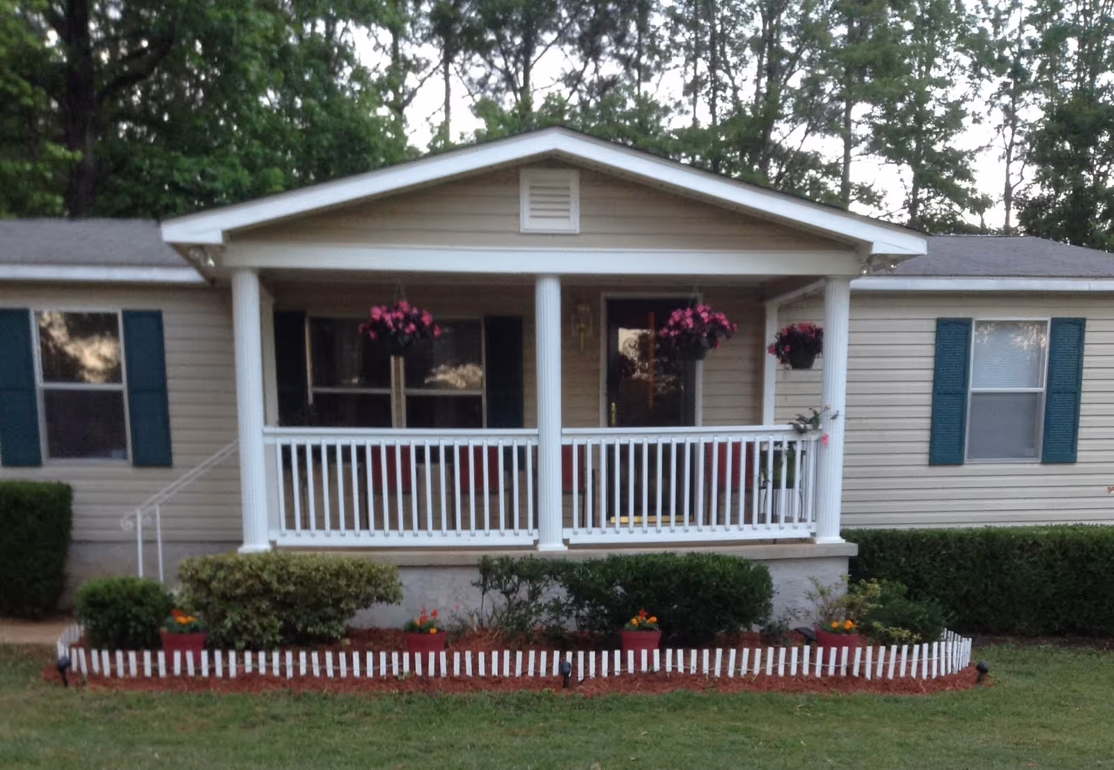 Front view of a single-story house with beige siding, a small porch with white railings and columns, hanging flower baskets, and a garden bed with shrubs and flowers bordered by a white picket fence.
