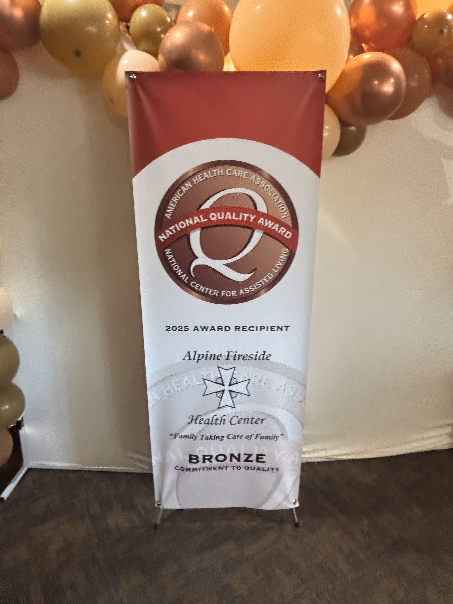A vertical indoor award banner for Alpine Fireside Health Center announcing the 2025 National Quality Award (Bronze) with decorative balloons above.