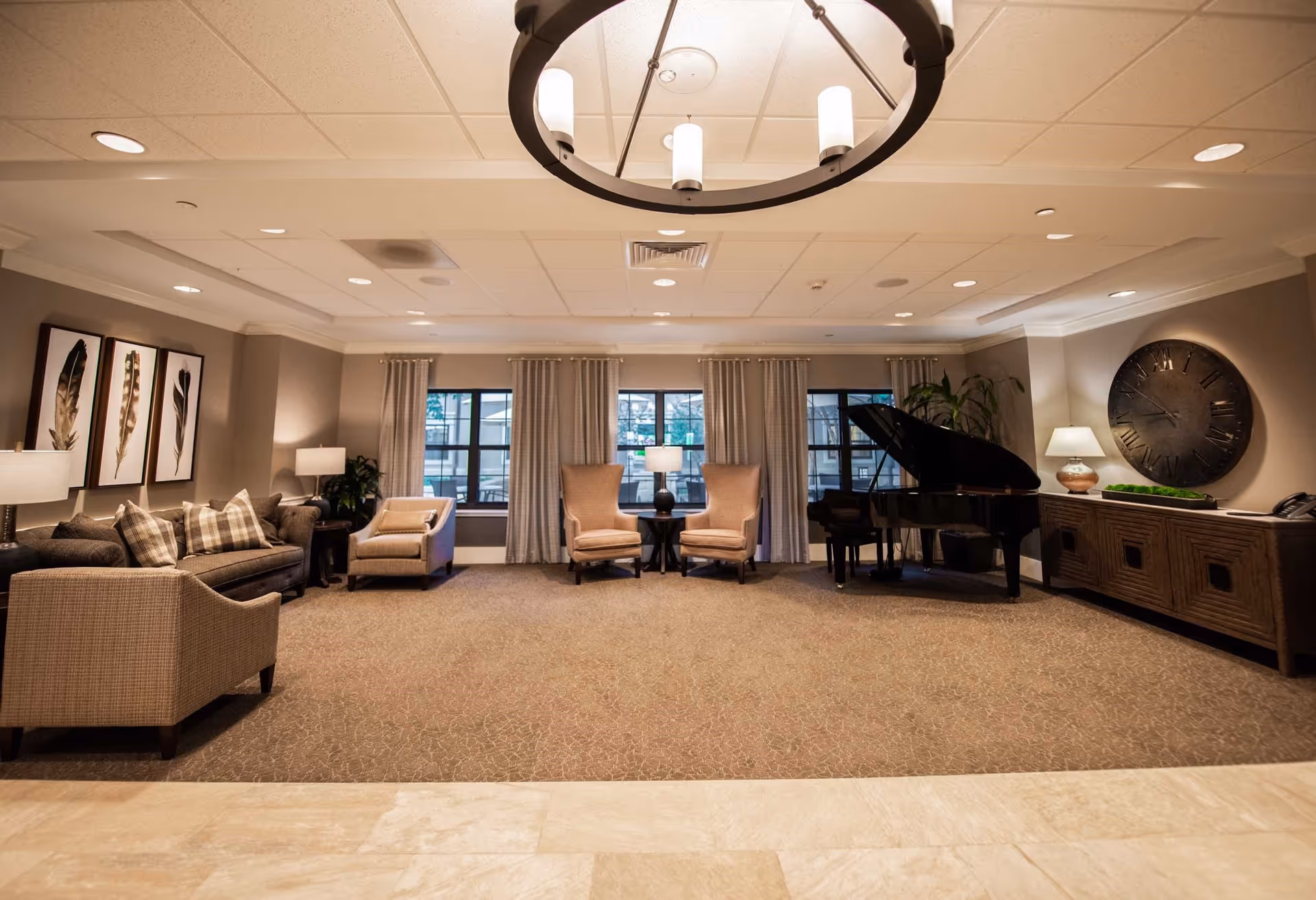 A spacious and well-lit senior living common area featuring a grand piano on the right, two beige armchairs with a small table and lamp between them in the center, and a sofa with patterned pillows and two armchairs on the left. The walls are decorated with framed feather artwork and a large round clock. The room has large windows with curtains and a modern circular ceiling light fixture.