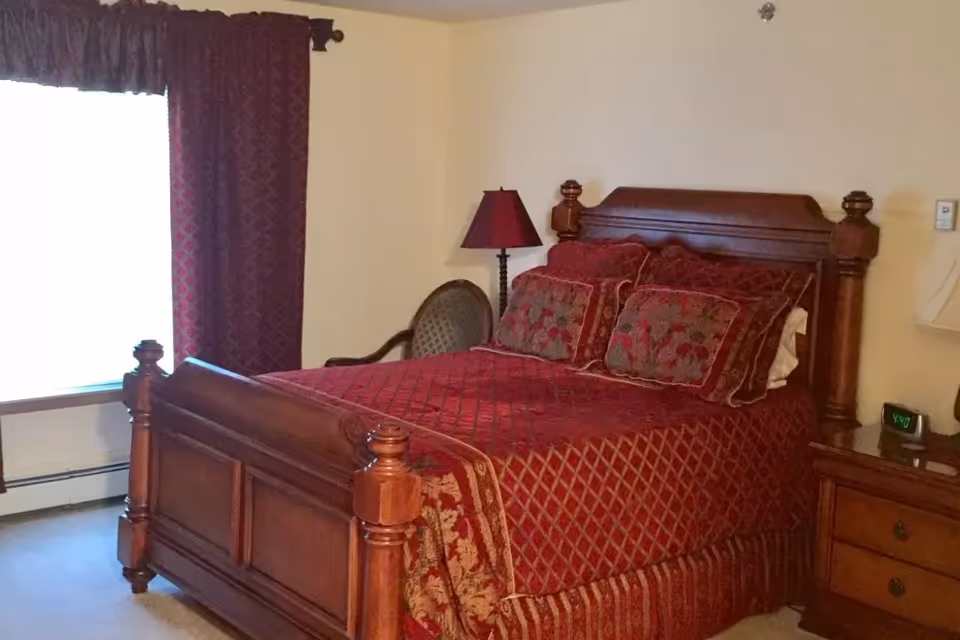 A bedroom with a wooden bed frame and headboard, covered with a red patterned bedspread and matching pillows. There is a wooden nightstand with a digital clock and a lamp, a window with red curtains, and a cushioned chair beside the bed.