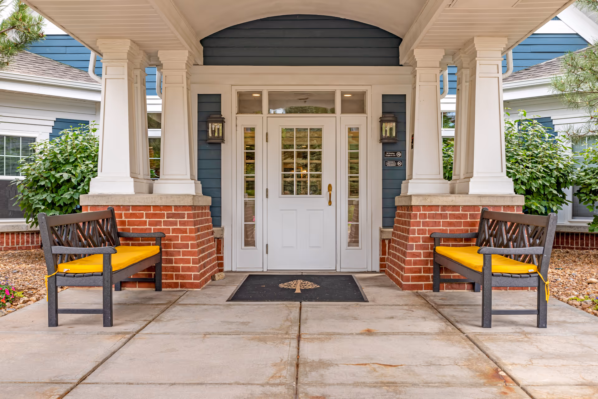 Covered entrance to a building with white double doors featuring glass panes, flanked by two white columns on brick bases. Two black benches with yellow cushions are placed symmetrically on either side of the entrance. Green shrubs and windows are visible on both sides of the entrance.