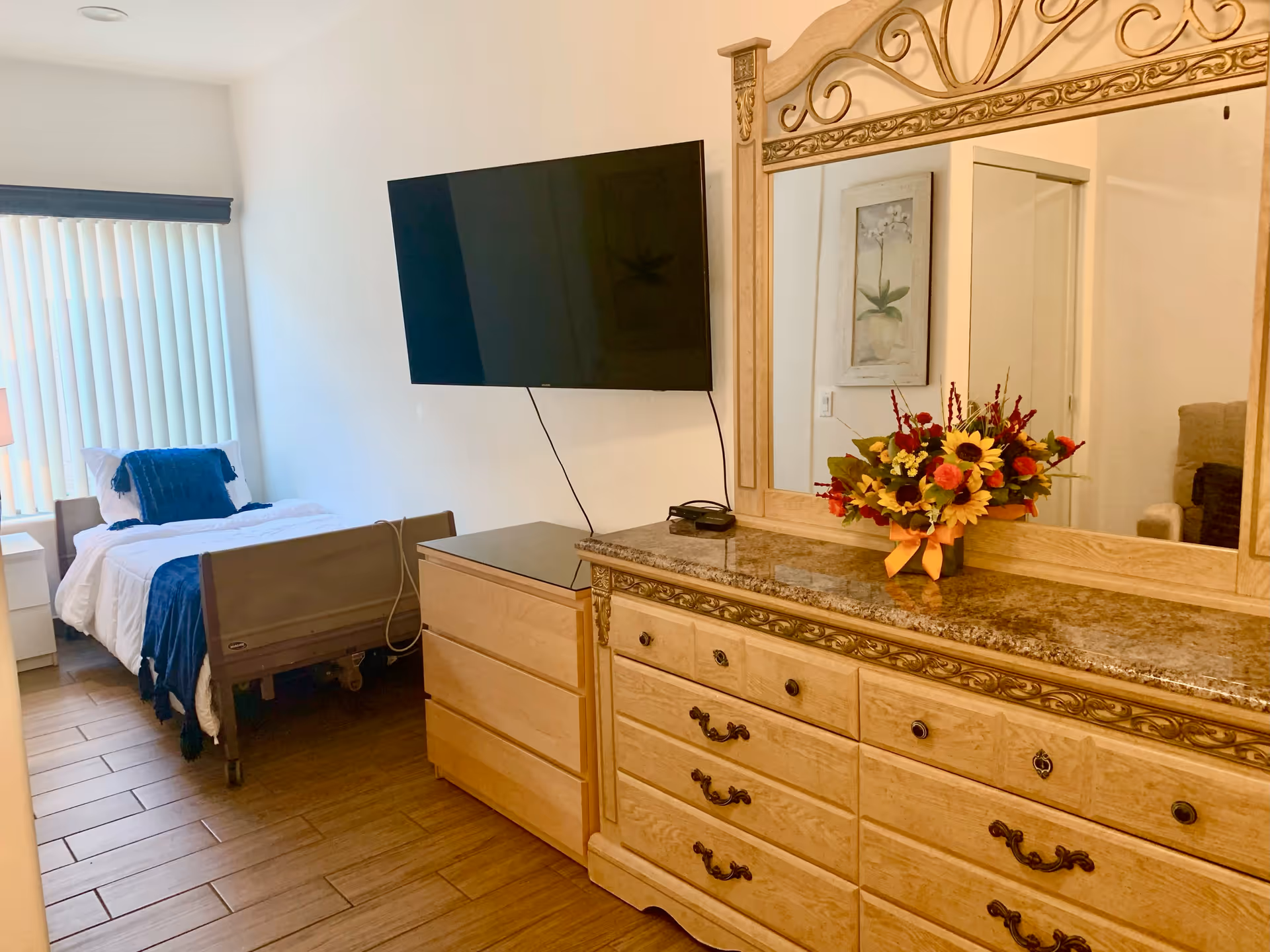 A furnished bedroom with an adjustable bed, wall-mounted TV, and a wooden dresser topped by a large mirror and a flower arrangement.