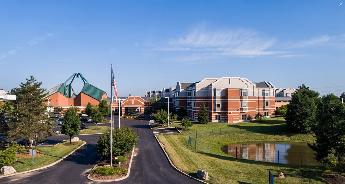 Exterior view of a large brick building with white trim, surrounded by green lawns, trees, a small pond, and a parking lot. An American flag is visible on a flagpole near the entrance driveway under a clear blue sky.