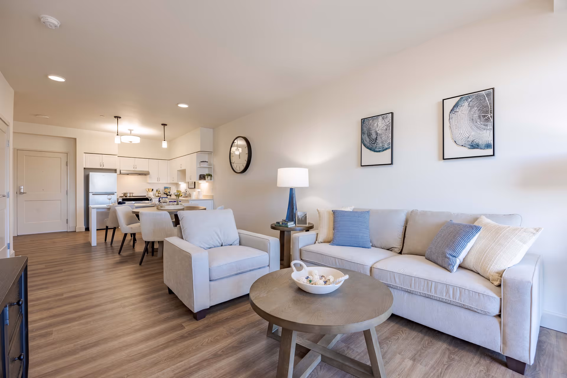 Open-plan living room with a beige sofa and armchair, round coffee table, and a view into the dining area and kitchen.