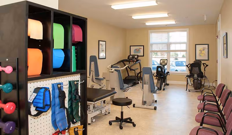 A well-lit exercise room with various fitness equipment including treadmills, stationary bikes, and weight machines. Colorful exercise mats and weighted vests are stored on a black shelving unit on the left. Several purple chairs are lined up against the right wall. The room has beige walls, a large window letting in natural light, and framed artwork on the walls.