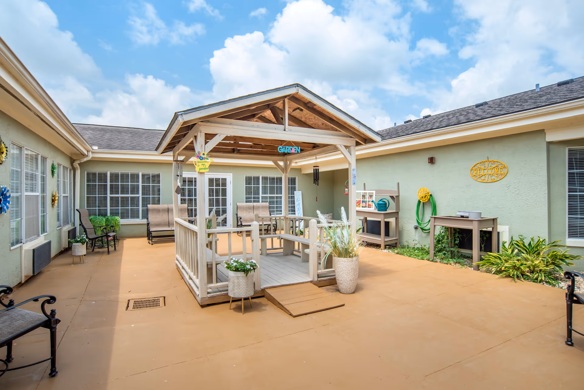 Enclosed courtyard patio featuring a wooden gazebo with seating, potted plants, and surrounding building windows under a blue sky.