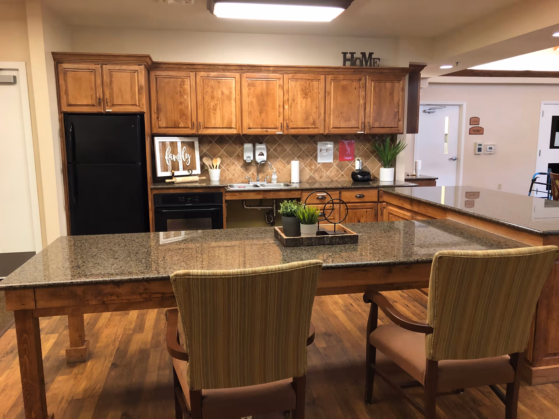 A kitchen area with wooden cabinets, a black refrigerator, and a black oven. There is a granite countertop island with two striped cushioned chairs. On the countertop, there is a decorative tray with small potted plants. The backsplash is tiled, and there are soap dispensers and a paper towel holder near the sink. The word 'HOME' is displayed on top of the cabinets.
