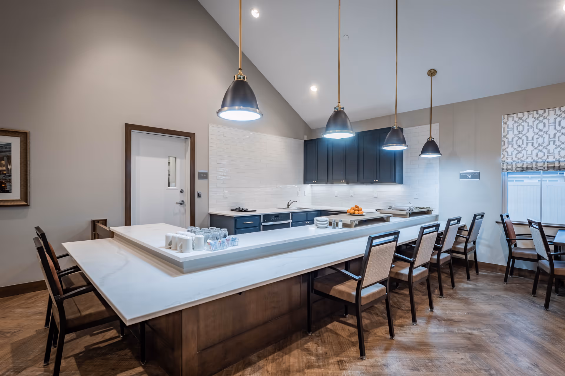 Modern kitchen area in a senior living facility with a large white marble countertop island surrounded by chairs. The kitchen features dark blue cabinets, white subway tile backsplash, and four pendant lights hanging from the ceiling. There are cups, glasses, and a bowl of oranges on the counter. A window with patterned blinds is visible on the right side.