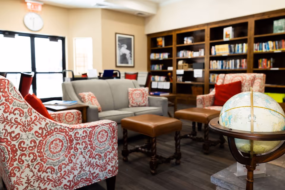 A cozy living room area in a senior living facility with patterned armchairs, a gray sofa with decorative pillows, two brown leather ottomans, a wooden globe stand with a globe, and a large bookshelf filled with books in the background.