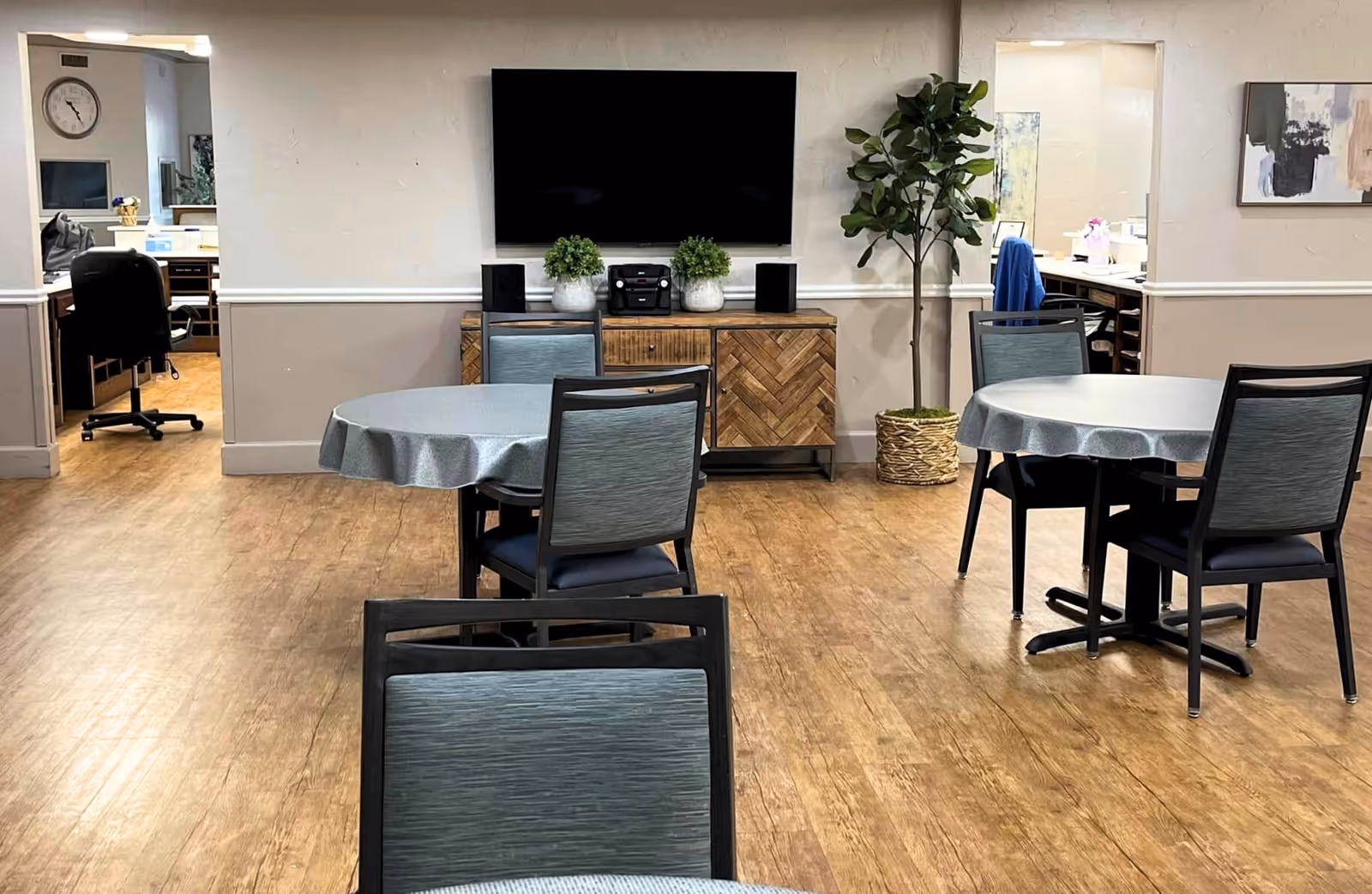 Interior view of a common area in a senior living facility with round tables covered with gray tablecloths and chairs around them. A large flat-screen TV is mounted on the wall above a wooden cabinet with two potted plants and a stereo system. There is a tall potted plant to the right of the cabinet. Two open doorways lead to office spaces with desks and chairs. The floor is wood-patterned, and the walls are painted in a two-tone style with a chair rail.