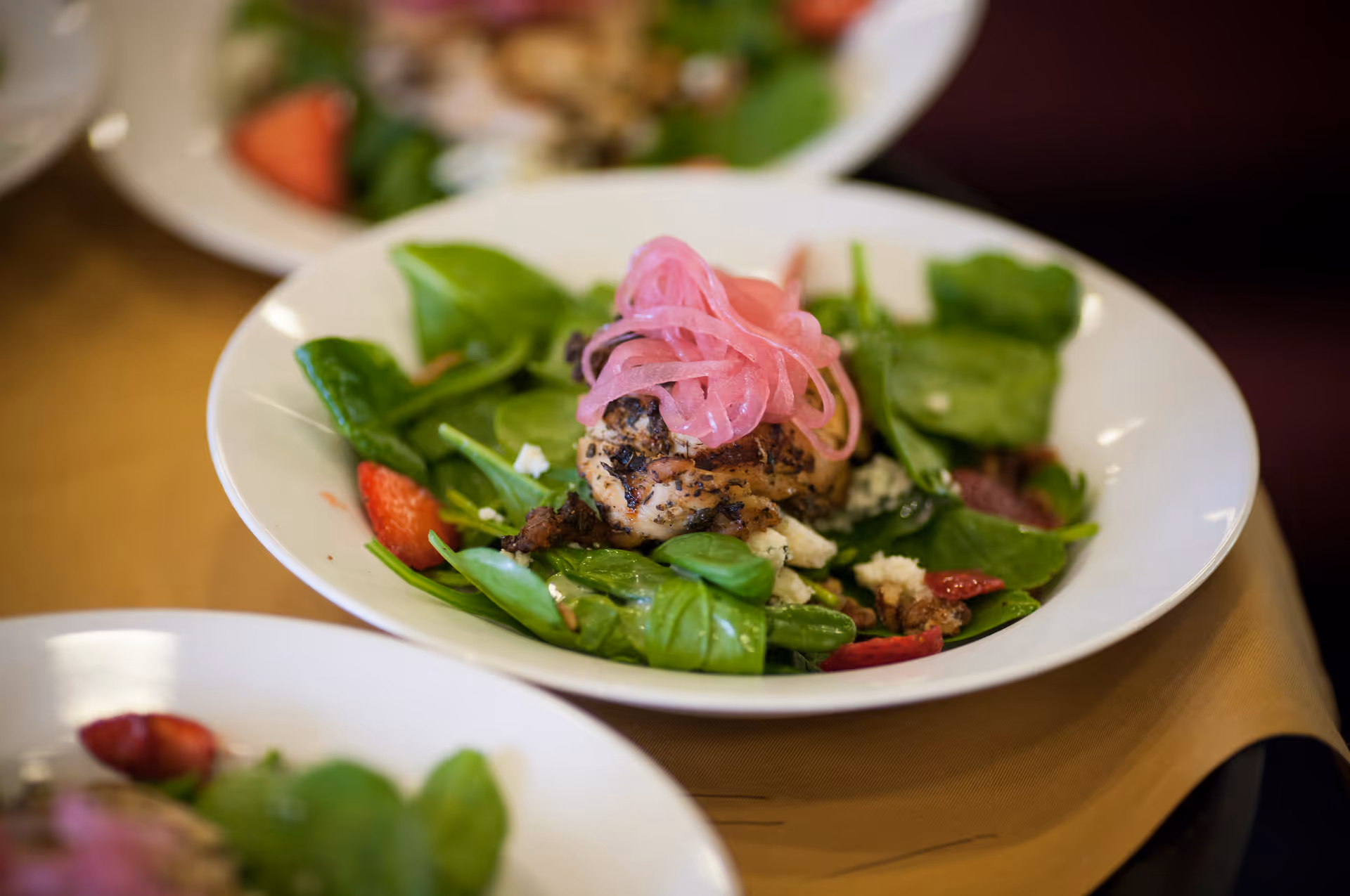 A close-up of a fresh salad served in a white bowl, featuring spinach leaves, grilled chicken, pickled red onions, strawberries, and crumbled cheese.