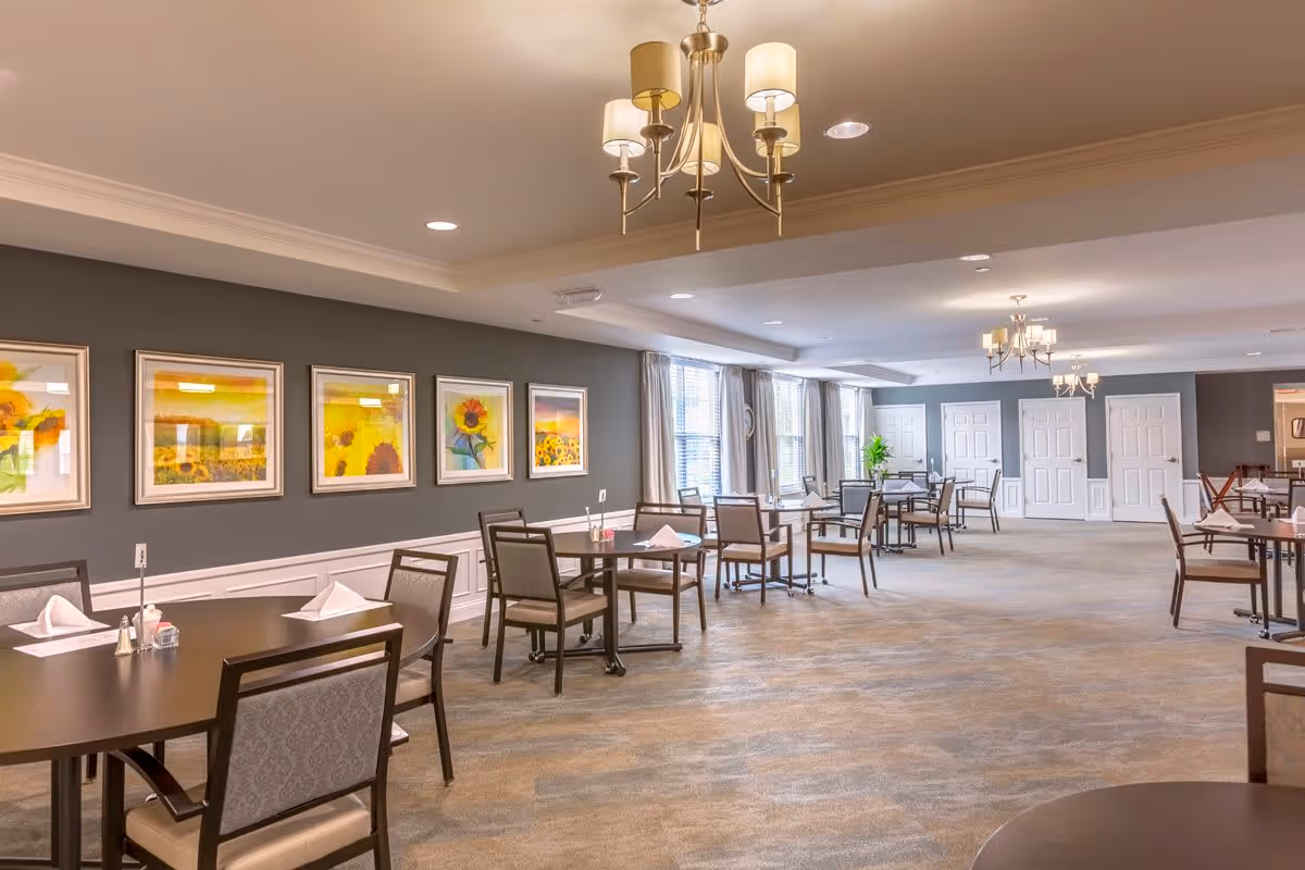 A spacious dining room with multiple round tables and chairs arranged neatly. The walls are decorated with framed sunflower-themed artwork. Large windows with curtains allow natural light to brighten the room. Elegant chandeliers hang from the ceiling, providing warm lighting.