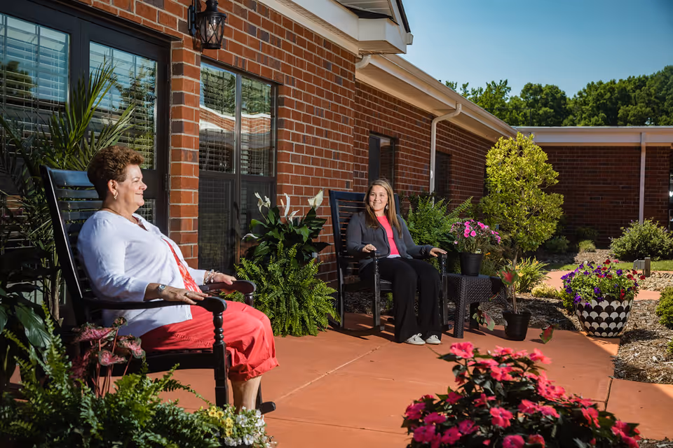 Two women sitting and relaxing on black rocking chairs on a patio outside a brick building, surrounded by various potted plants and flowers under a clear sky.