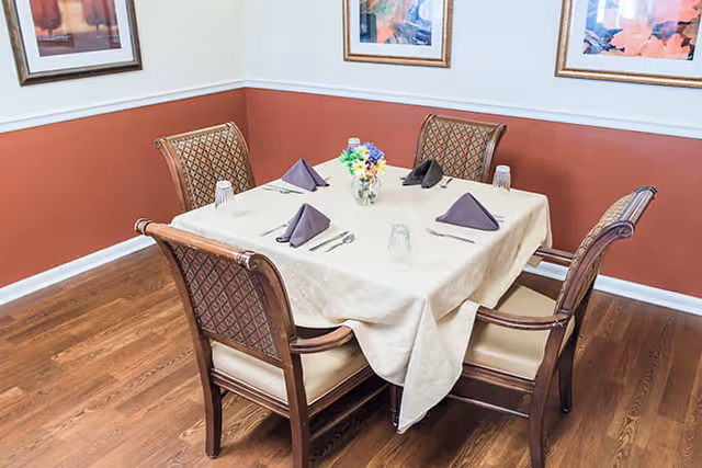 Square dining table set for four with folded napkins and a small floral centerpiece in a corner dining room.