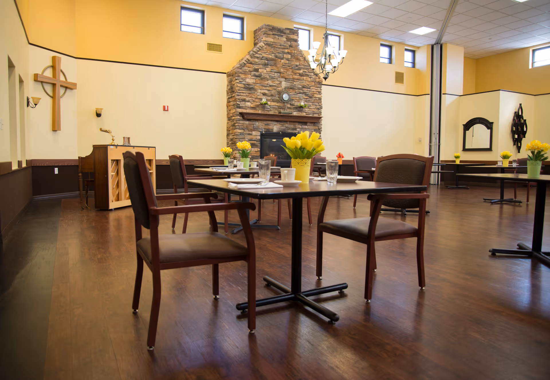 A spacious dining room with wooden floors and several tables set with glasses, cups, and yellow flower centerpieces. The room features a large stone fireplace, a chandelier, a piano, and a wooden cross mounted on the wall. The walls are painted in light yellow and brown tones, with high windows letting in natural light.