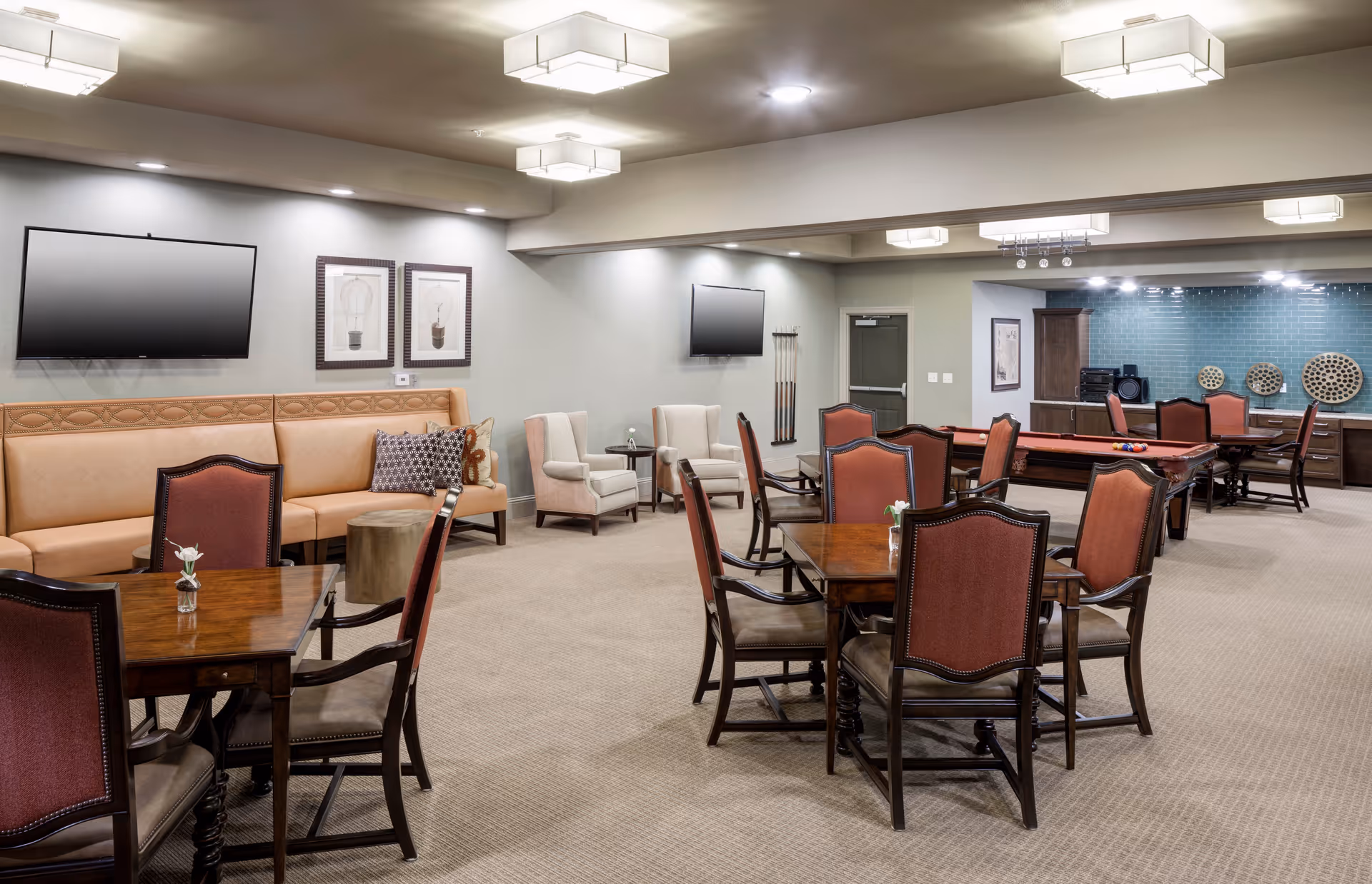 A spacious senior living common area with multiple wooden tables and chairs, a tan leather bench along one wall with decorative pillows, two beige armchairs, two wall-mounted flat-screen TVs, a pool table with billiard balls, and a game area with wooden game boards on a teal tiled wall. The room is well-lit with ceiling lights and has a carpeted floor.