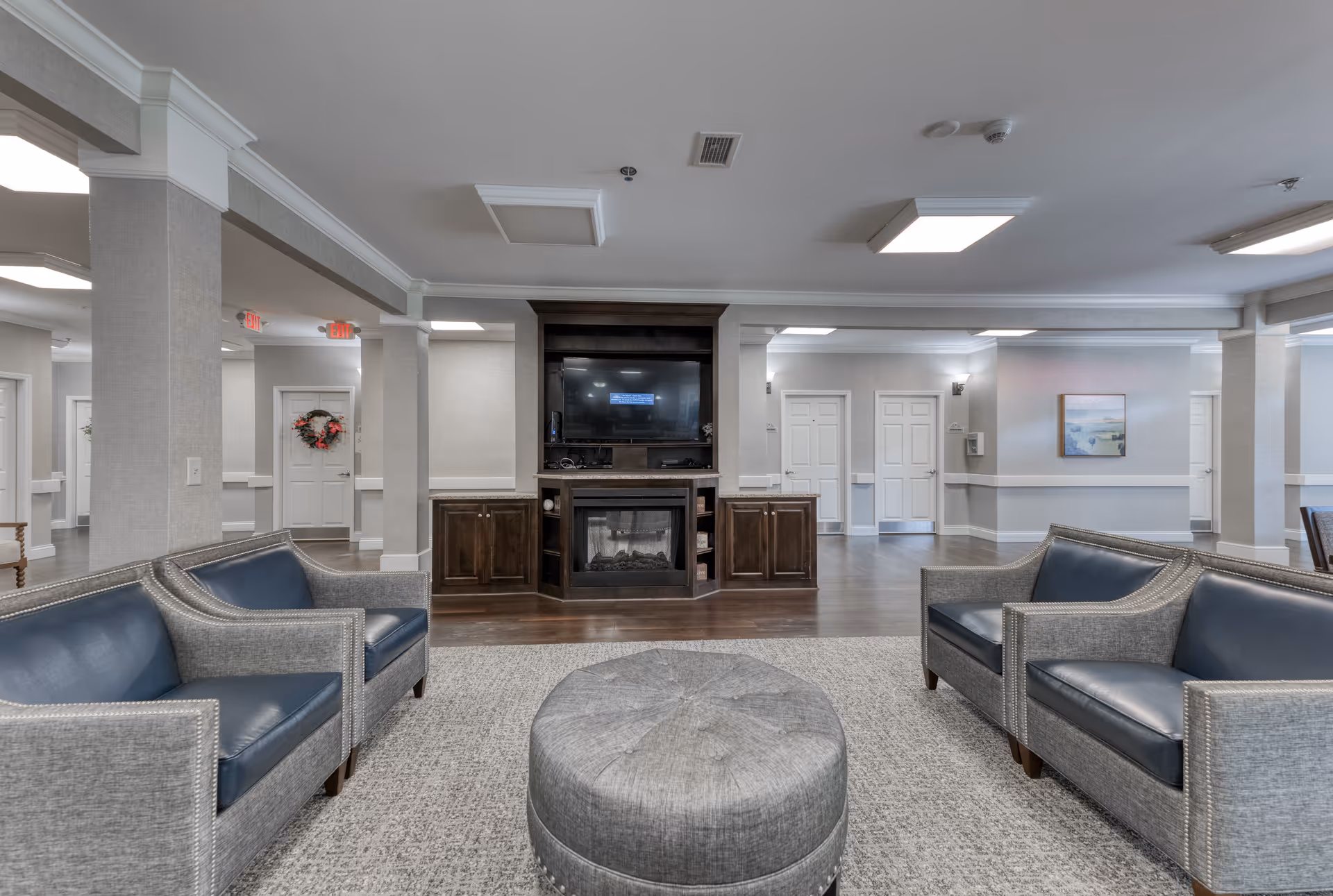 A spacious senior living facility common area with four gray armchairs featuring blue cushions arranged around a round gray ottoman on a textured carpet. In the background, there is a dark wood entertainment center with a flat-screen TV mounted above an electric fireplace. The walls are light gray with white trim, and there are multiple white doors and columns visible, along with ceiling lights providing bright illumination.