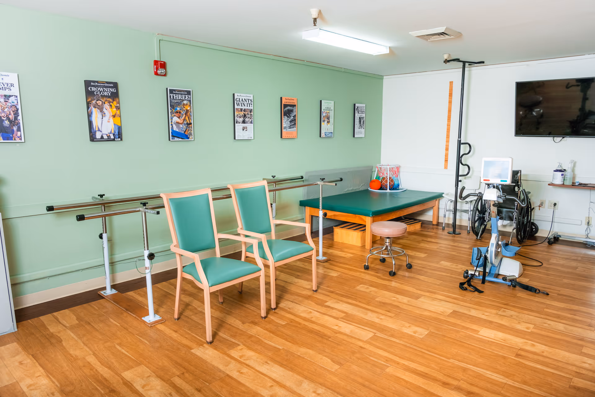 A physical therapy room with wooden flooring, two green padded chairs, parallel bars for walking exercises, a green padded therapy table, a stool on wheels, a wheelchair, an exercise bike, and a wall-mounted TV. The walls are light green with framed sports-themed posters.
