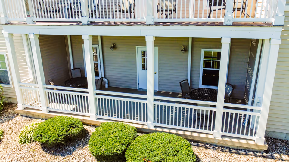 Covered outdoor patio area of a senior living facility with white railings and columns, two round metal tables with chairs, beige siding walls, a white door, and windows. Green bushes and gravel landscaping are visible in front of the patio.