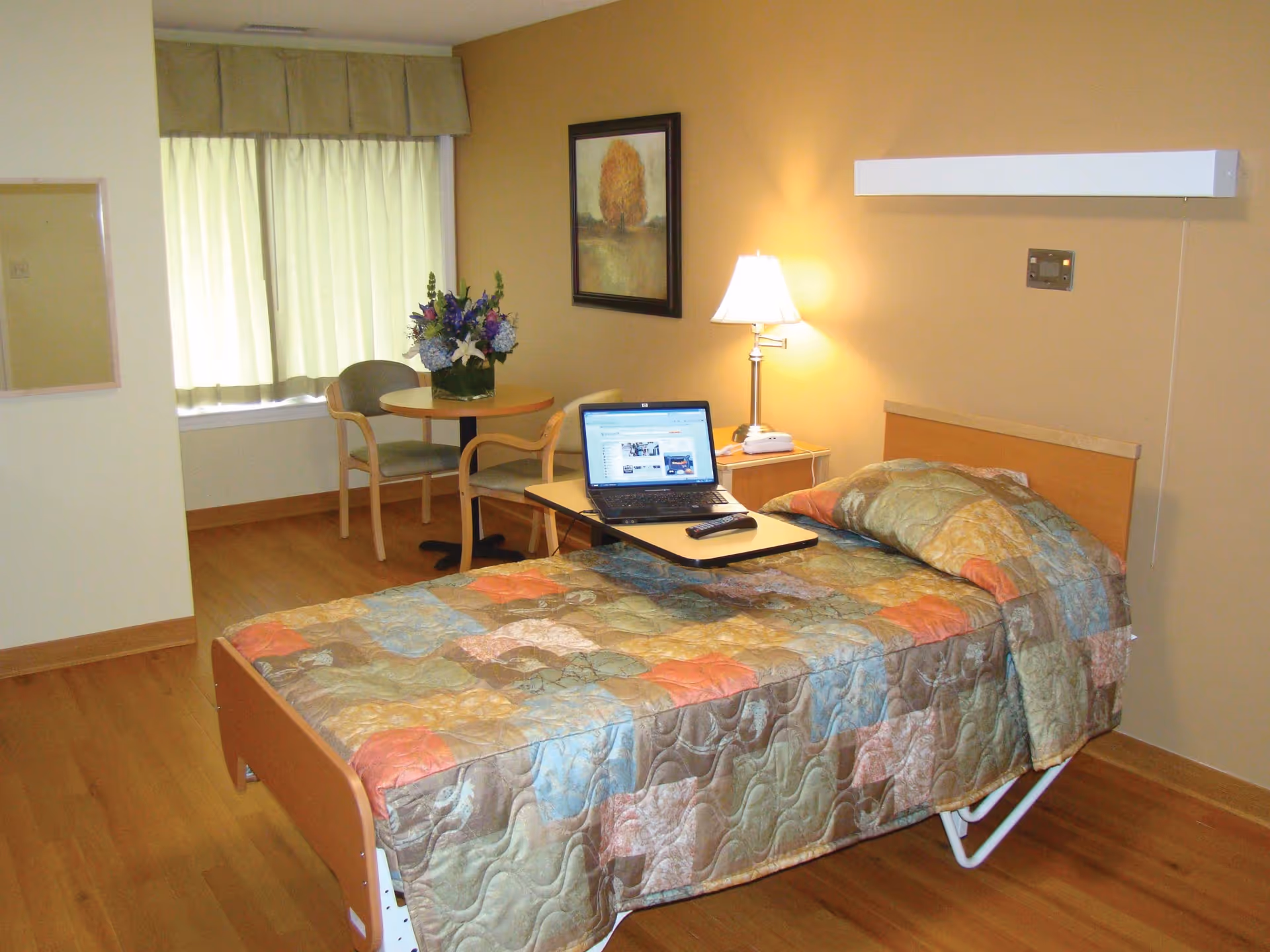 A single bed with a colorful patchwork quilt in a senior living facility room. A laptop and remote control are on an overbed table. There is a small round table with two chairs and a vase of flowers near a window with closed curtains. A framed picture hangs on the wall above a bedside table with a lamp and a phone.