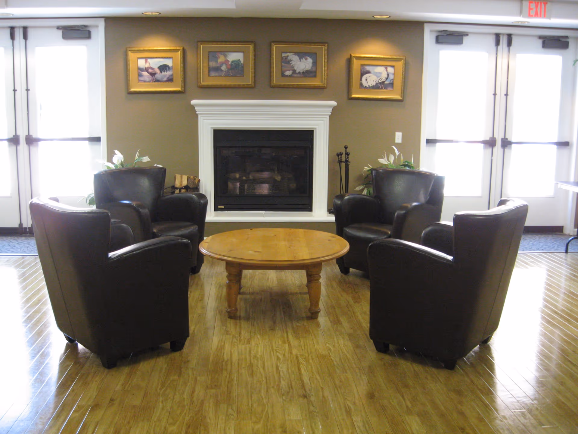 A cozy seating area with four dark leather armchairs arranged around a round wooden coffee table in front of a white-framed fireplace. Above the fireplace are four framed pictures of roosters. There are double glass doors on either side of the fireplace, and plants are placed near the chairs.