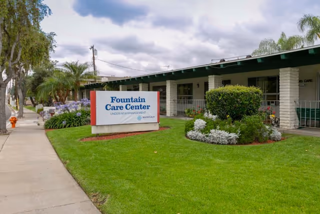 Exterior view of Fountain Care Center building with a green lawn, bushes, and flowers in front. A sidewalk runs alongside the building and a sign in the grass reads 'Fountain Care Center Under New Management'. The sky is cloudy.
