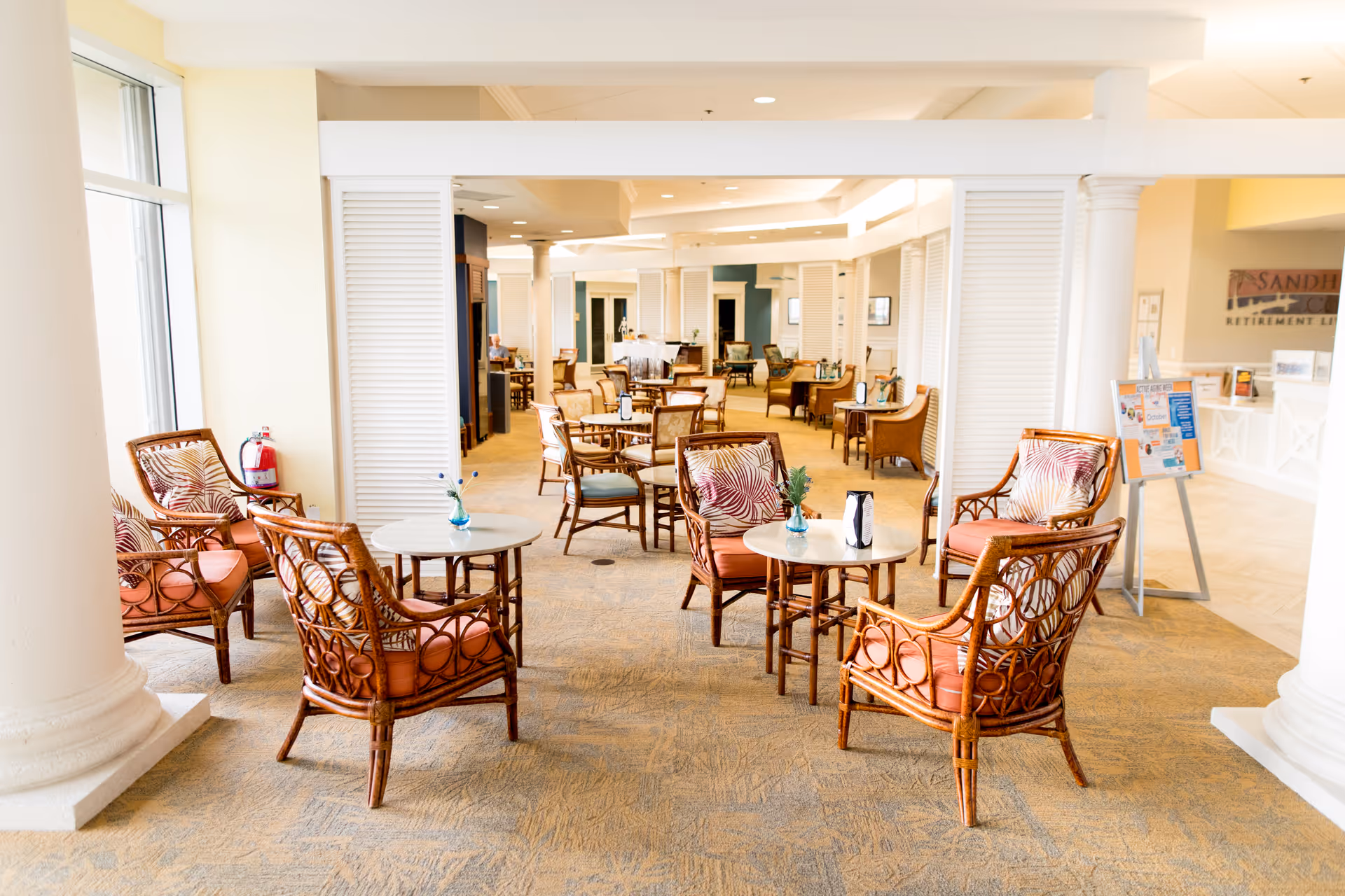 A bright and spacious common area in a retirement living facility with multiple round tables surrounded by wicker chairs with cushions. The room has large windows letting in natural light, white columns, and a carpeted floor. In the background, more seating and tables are visible along with a reception desk and a sign that reads Sandhill Cove Retirement Living.