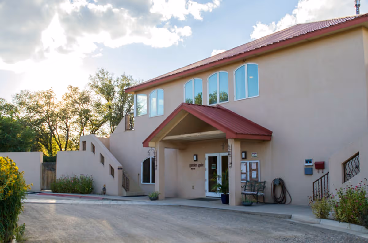 Exterior view of a two-story beige building with a red roof and an entrance covered by a small red awning. There are stairs on both sides of the building, several windows, and some greenery around the entrance area. The sky is partly cloudy with sunlight filtering through.