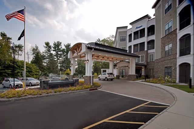 Entrance of Allegro Senior Living facility in Harrington Park, NJ, showing a covered driveway with a white car parked underneath. The building is multi-story with balconies, and there is a fountain and American flag on the left side near the parking area.