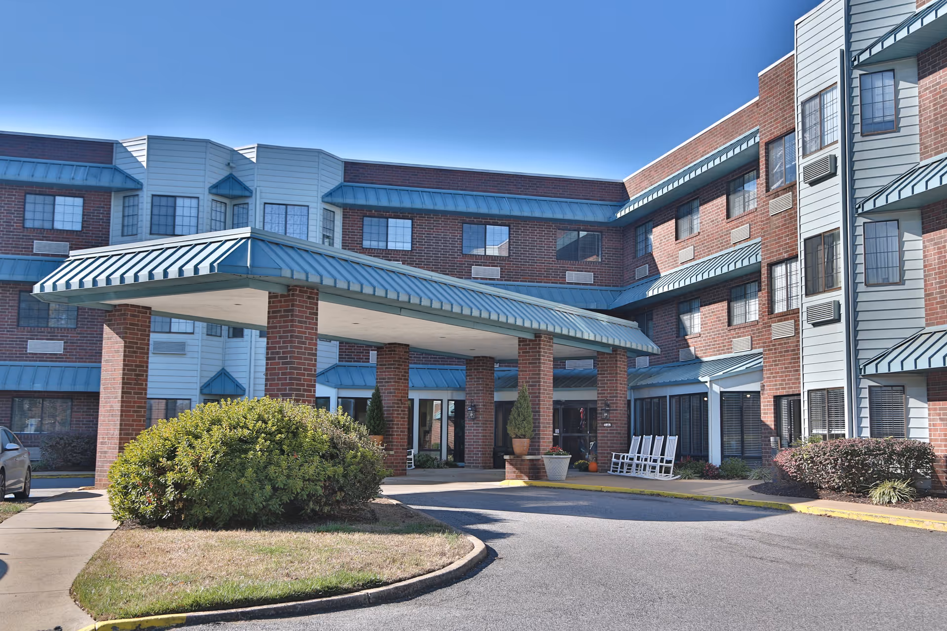 Exterior view of a multi-story senior living facility building with a covered entrance supported by brick columns. The building features red brick and light blue siding with multiple windows and blue awnings. There are bushes and a small lawn area near the entrance, along with a few white rocking chairs on the right side.