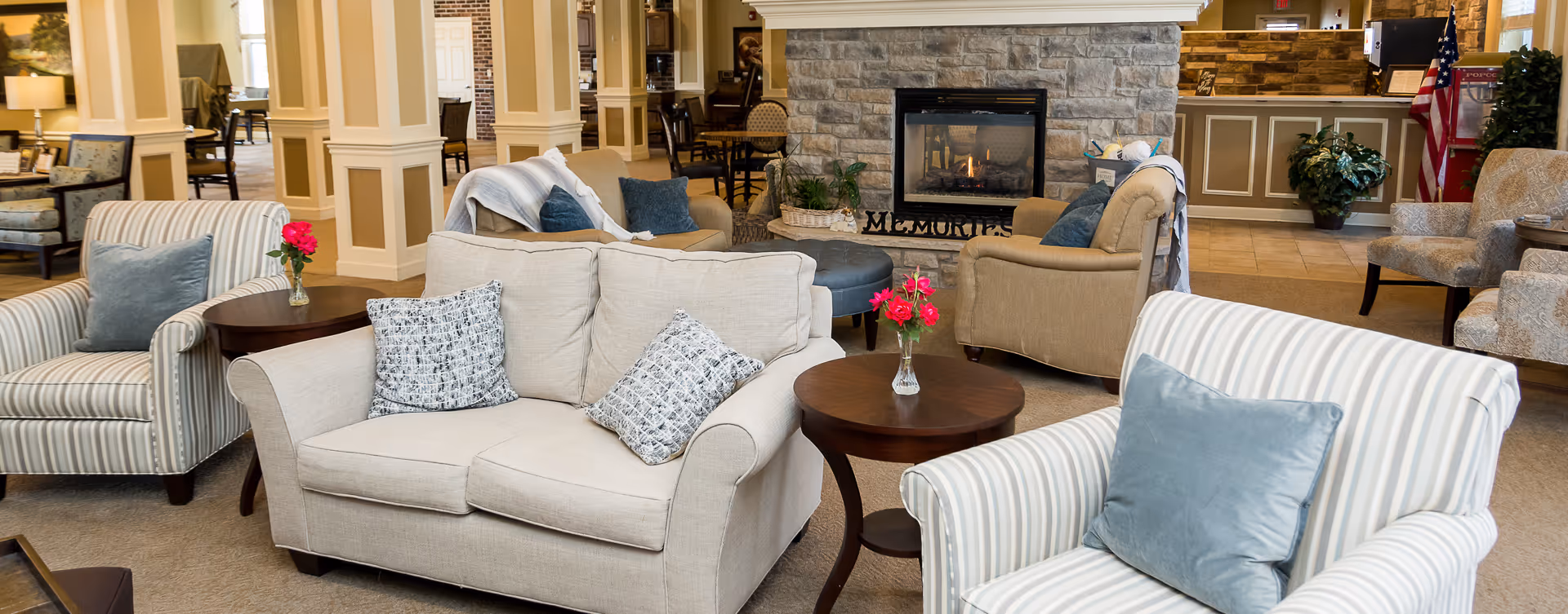 A cozy senior living facility common area with beige and striped armchairs, a beige loveseat with patterned pillows, wooden side tables with small vases of red flowers, and a stone fireplace with a fire burning. The background shows more seating, plants, and a reception desk with an American flag nearby.