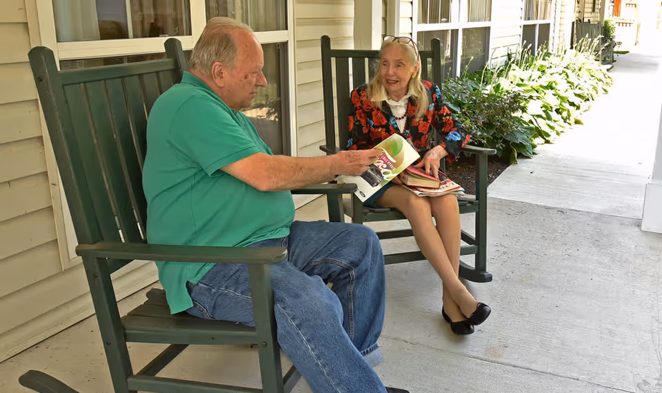 An elderly man and woman sitting on green rocking chairs on a covered porch outside a building. The man is wearing a green polo shirt and jeans, holding a book, while the woman, dressed in a floral jacket and black shoes, is looking at him and smiling. There are plants along the side of the porch and windows behind them.