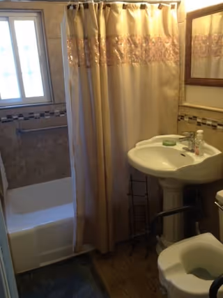 Small bathroom with a white bathtub partially covered by a beige shower curtain with a decorative band. There is a white pedestal sink with a soap dish and a bottle of hand soap on it. A mirror with a wooden frame hangs above the sink. The bathroom has tiled walls and a window with frosted glass on the left side. A toilet with a raised seat and support bars is visible in the foreground.