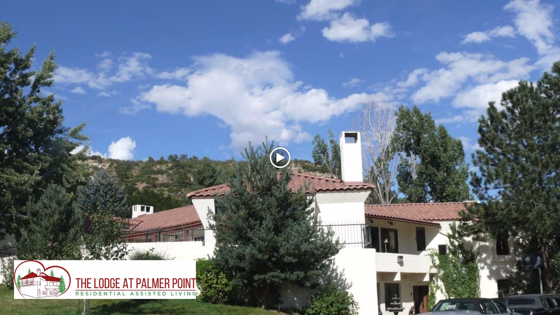 Exterior view of a white two-story assisted living building with red tile roof surrounded by trees under a blue sky.
