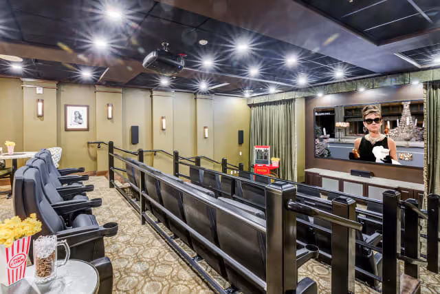A small theater room with multiple rows of black leather seats facing a large screen showing a scene from the movie Breakfast at Tiffany's. The room has beige walls with framed pictures, wall sconces, and a patterned carpet. There is a popcorn machine and popcorn containers visible.