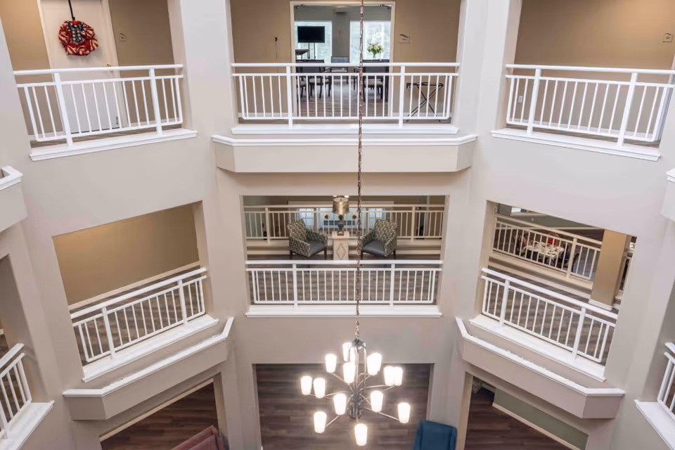 Interior view of a multi-level atrium in a senior living facility with white railings on each floor. There are seating areas with chairs and tables visible on the middle floors, and a modern chandelier hanging in the center. The walls are painted beige, and the floors have a wood-like finish.